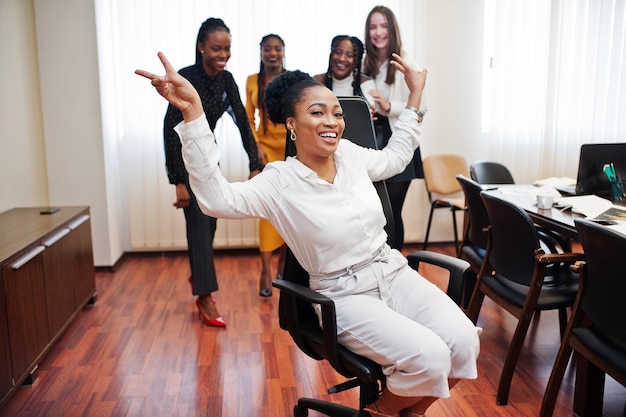 Cinco mujeres de negocios multirraciales de pie en la oficina y ruedan a la mujer en la silla Diverso grupo de empleadas en ropa formal divirtiéndose