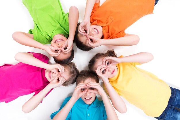 Cinco niños felices tirados en el suelo en círculo con las manos cerca de los ojos en camisetas brillantes. Vista superior. Aislado en blanco.