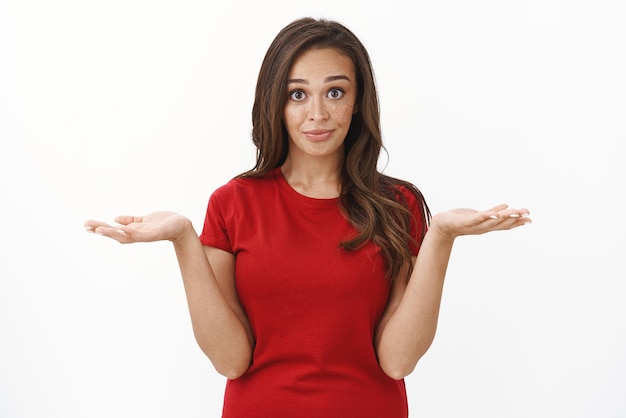 Cintura arriba disparó a una linda mujer morena que se enfrenta a una elección difícil encogiéndose de hombros y tomándose de la mano mientras elige entre dos variantes sonríe pidiendo consejo de pie con fondo blanco en una camiseta roja Copiar espacio