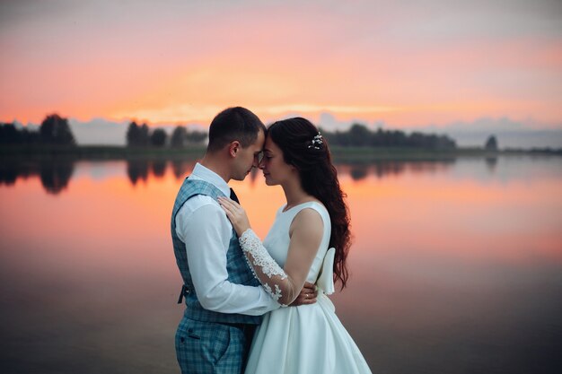 Cintura para arriba de la pareja de novios románticos abrazándose y posando junto al lago al atardecer con una vista increíble. Novios en concepto de amor
