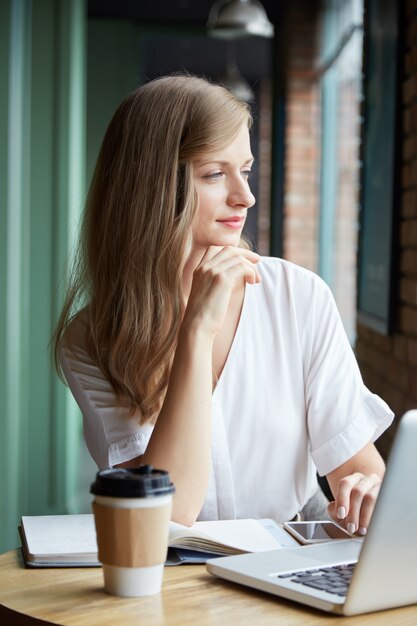 Cintura para arriba de pensativa joven sentada en el escritorio mirando por la ventana