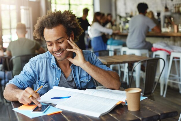 Cintura para arriba retrato de estudiante universitario afroamericano sonriente positivo escribiendo un mensaje de texto en su teléfono inteligente con pantalla en blanco de espacio de copia mientras está sentado en la cantina y trabajando en una tarea en casa