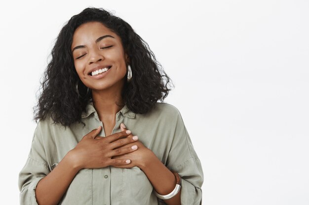 Cintura para arriba de la tierna niña afroamericana soñadora y conmovida con peinado rizado sosteniendo palmas en el corazón cerrando los ojos y sonriendo nostálgico