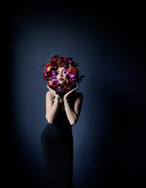 Círculo colorido hecho de flores frescas en la cara de la hermosa niña, mujer vestida con un vestido negro ajustado sobre el fondo azul oscuro