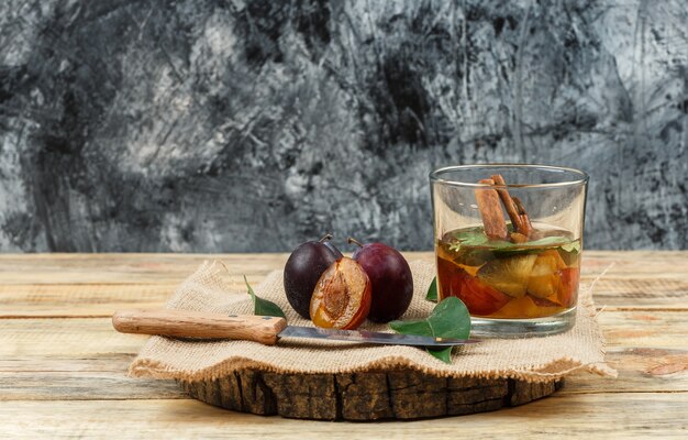 Ciruelas de primer plano y cuchillo sobre tablero de madera con agua de desintoxicación, un pedazo de saco y hojas sobre tablero de madera y superficie de mármol azul oscuro. horizontal