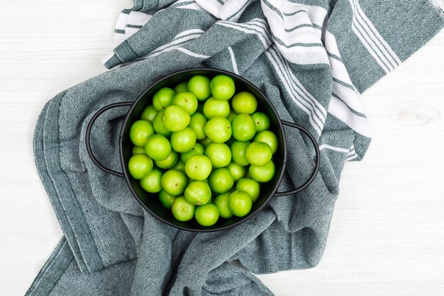 Ciruelas verdes en una cacerola negra sobre papel de cocina y madera blanca. vista superior.