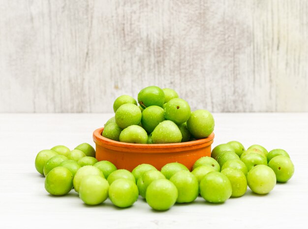 Ciruelas verdes cubiertas de rocio en un recipiente de arcilla redonda en blanco y grunge, vista lateral.