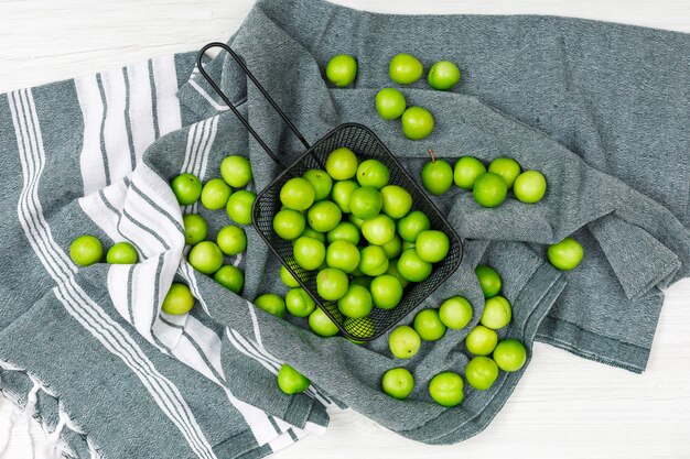 Ciruelas verdes dispersas en un cullender negro sobre una toalla de cocina y madera blanca, vista desde arriba.