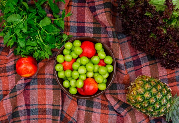 Ciruelas verdes y duraznos en un tazón de arcilla con hojas verdes, una piña y una lechuga planas sobre una tela de picnic