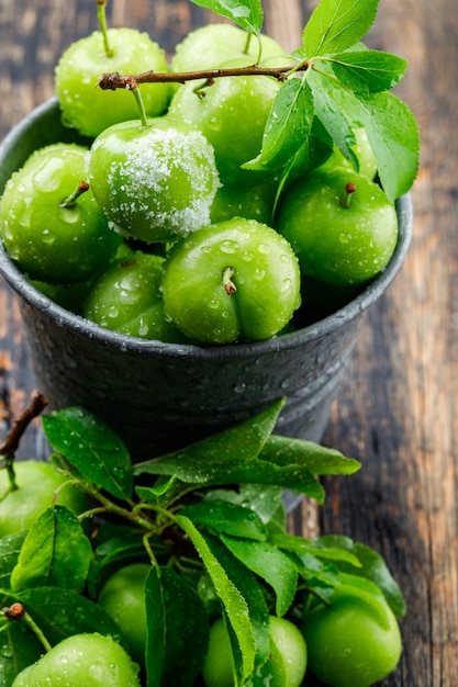 Ciruelas verdes saladas en un mini cubo con hojas de alto ángulo de visión en una pared de madera