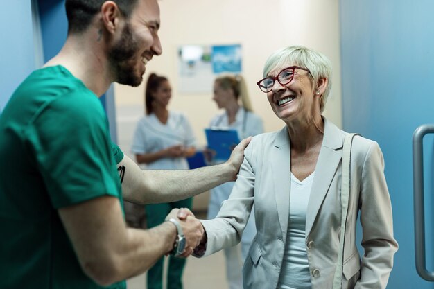 Cirujano feliz y anciana estrechando la mano mientras saludan en el vestíbulo de la clínica El foco está en la mujer