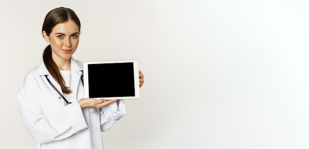Foto gratuita cita remota de ayuda médica en línea hermosa mujer doctora sonriente que muestra la pantalla de la tableta digital que muestra el logotipo del sitio web de pie sobre fondo blanco