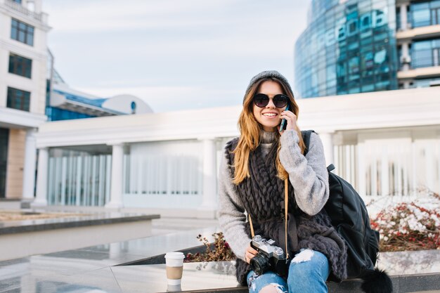 Ciudad con estilo alegre joven sentada en el centro de la ciudad moderna de Europa, vistiendo suéter de lana de invierno, gafas de sol, gorro de punto. Hablando por teléfono, viajando con bolsa, cámara, sonriendo. Lugar para el texto.