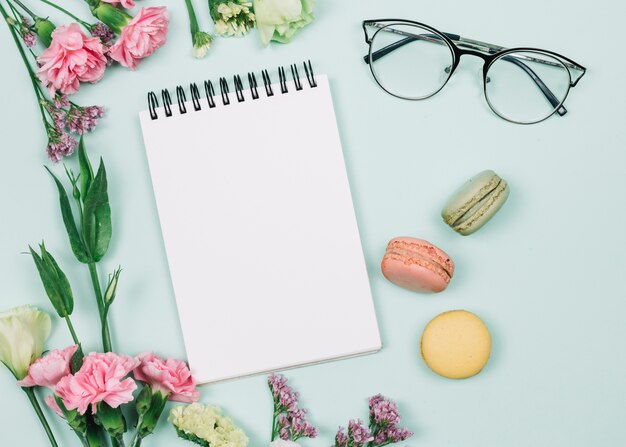 Claveles rosados y flores de limonium cerca de la libreta espiral; anteojos y macarrones sobre fondo azul