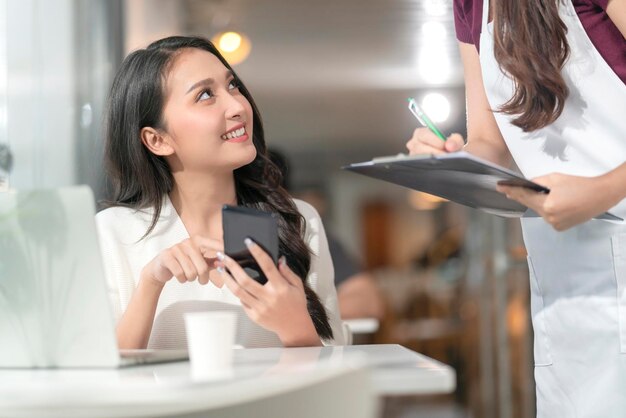 Una clienta asiática pide comida y bebida del menú digital del teléfono inteligente social sin contacto al personal de camarera Camarera milenaria sonriente con un gráfico de papel tomando el pedido de una joven clienta asiática