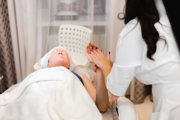 Una clienta joven con una máscara de alginato en su rostro se encuentra en la mesa de cosmetología, la esteticista hace un masaje manual de manos