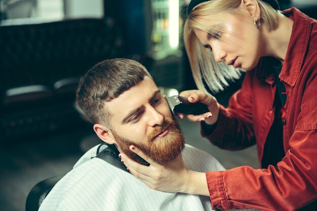 Cliente durante el afeitado de barba en peluquería. Peluquería femenina en el salón. Igualdad de género. Mujer en la profesión masculina.