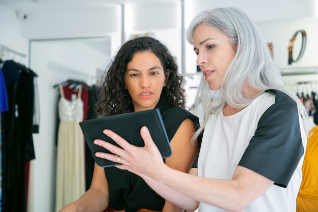 Cliente y asistente de tienda reunidos en una tienda de moda, sentados juntos y usando la tableta, discutiendo sobre ropa y compras. Consumismo o concepto de compras