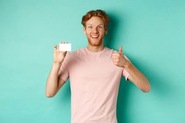 Cliente de banco masculino alegre en camiseta mostrando el pulgar hacia arriba y tarjeta de crédito plástica, sonriendo satisfecho a la cámara, de pie sobre fondo turquesa.