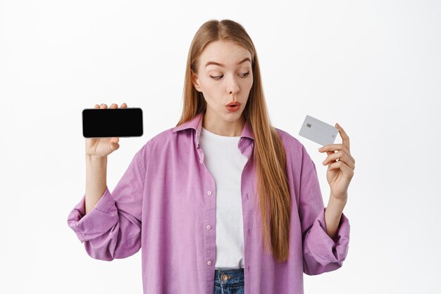 Cliente de banco de mujer joven mirando asombrado por la tarjeta de crédito y mostrando la pantalla del teléfono inteligente de la aplicación de teléfono móvil volteada de pie horizontal sobre fondo blanco