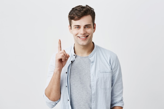 Cliente caucásico satisfecho y satisfecho señalando con el dedo índice hacia arriba en el espacio en blanco encima de su anuncio. Apuesto hombre positivo sonriendo con dientes y gesticulando, posando en el estudio