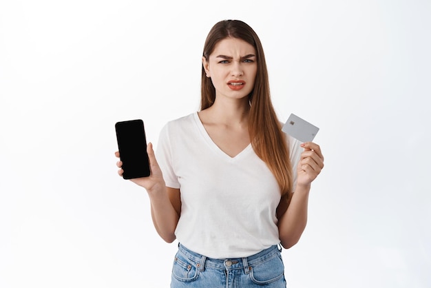 Cliente femenino decepcionado con el ceño fruncido que muestra el teléfono inteligente y la tarjeta de crédito se ven frustrados expresando disgusto quejándose en la aplicación de compras en línea de pie disgustado contra el fondo blanco