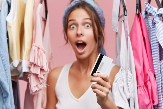 Cliente femenino parado en una boutique cerca de un estante con ropa, sorprendiéndose con los altos precios, sosteniendo una tarjeta de plástico en la mano mientras tiene poco dinero en su cuenta, queriendo comprar un vestido nuevo