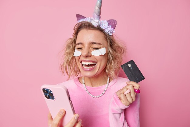 Clientes bancarios Mujer hermosa positiva sostiene tarjeta de crédito y teléfono inteligente se ríe alegremente feliz de ganar dinero se somete a procedimientos de belleza usa vestido de diadema de unicornio y collar aislado en rosa
