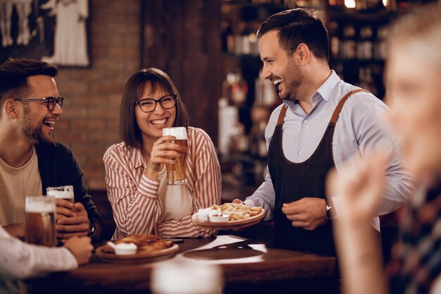 Clientes felices bebiendo cerveza y divirtiéndose con un camarero que les sirve comida en una taberna