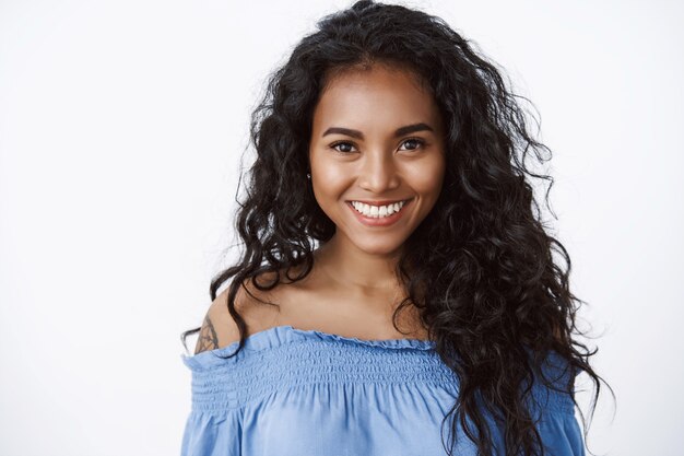 Close-up atractiva joven confiada y carismática con cabello largo y rizado, usar blusa azul, sonrisa dentuda y mirada sincera con expresión segura de sí misma y alentada