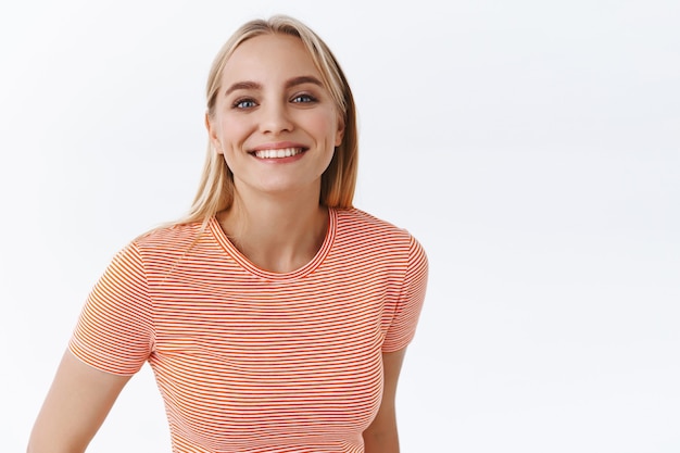 Close-up atractiva mujer caucásica rubia femenina animada en camiseta a rayas, sonriendo con dientes y entusiasta, sonrisa de alegría y felicidad, de pie fondo blanco optimista y alegre