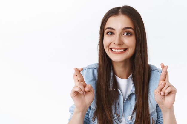 Close-up esperanzada, optimista linda chica morena cree que los sueños se hacen realidad, tienen aspiraciones y sueños, cruzan los dedos para la buena suerte, sonríen, rezan y anticipan buenas noticias, fondo blanco