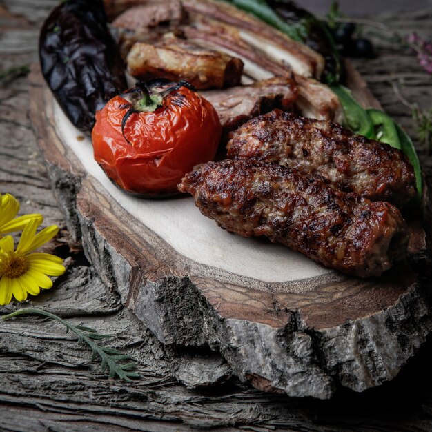 Close-up kebab costillas a la parrilla lula kebab con tomate al horno, pimiento y berenjenas en un plato de madera sobre una corteza de madera oscura