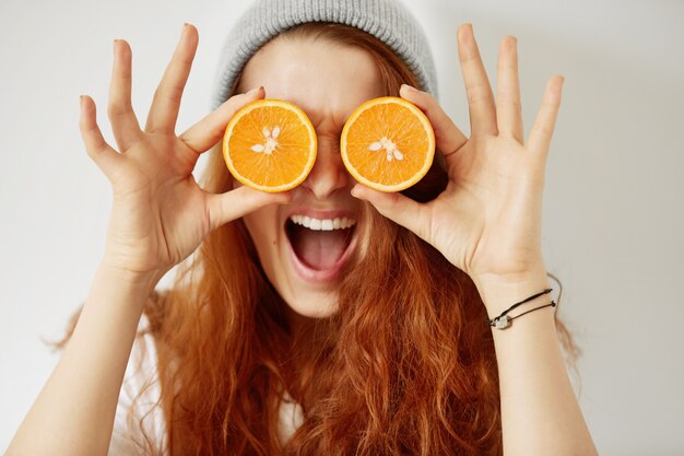Close up retrato aislado de mujer joven pelirroja sosteniendo naranjas a la mitad en sus ojos