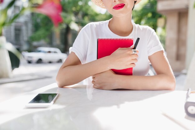 Close-up retrato al aire libre de dama romántica en camisa blanca con diario rojo y sonriendo