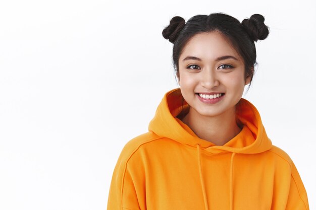Close-up retrato amigable feliz chica asiática kawaii en sudadera con capucha naranja con dos lindos moños, sonriendo feliz, muestra emociones positivas y alegres, pared blanca de pie, concepto de belleza