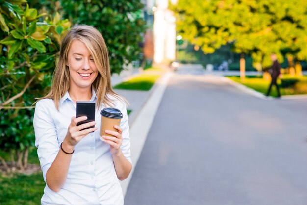 Close up retrato de una empresaria alegre utilizando su teléfono inteligente y carpetas de trabajo de transporte, de pie contra un cielo azul soleado.