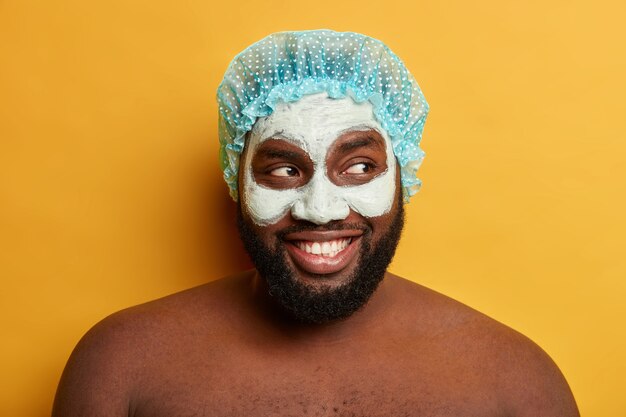 Close Up retrato de feliz chico de piel oscura lleva gorro de baño, aplica máscara de arcilla cosmética