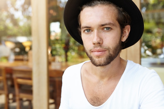 Close Up retrato de guapo joven inconformista con barba difusa con sombrero negro y camiseta blanca mirando con una leve sonrisa