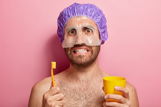 Close Up retrato de hombre guapo sostiene cepillo de dientes y taza amarilla, se para con shouders desnudos, tiene máscara en la cara