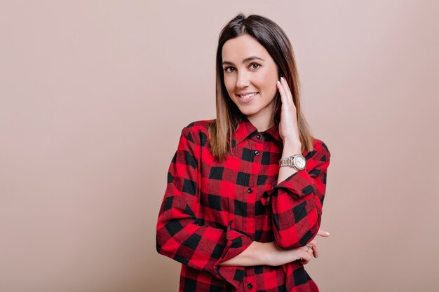 Close-up retrato de joven encantadora vestida con camisa a cuadros sonríe en el frente y sostiene la mano cerca de la cara