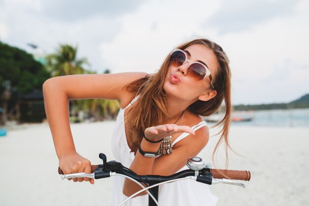 Close Up retrato de joven sonriente en vestido blanco montando en playa tropical en gafas de sol de bicicleta viajando en vacaciones de verano en Tailandia