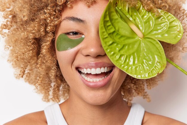 Close up retrato de mujer alegre y alegre sonríe ampliamente tiene dientes blancos perfectos cubre los ojos con lirio de cala verde aplica parches de hidrogel para reducir las poses de arrugas en interiores. Concepto de belleza y mujeres