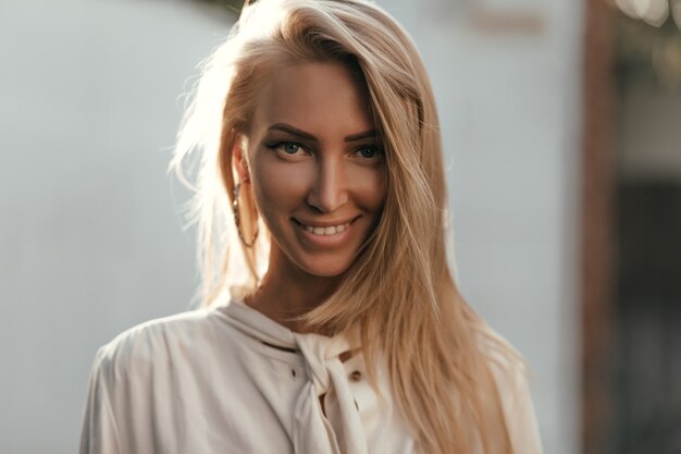 Close-up retrato de mujer bronceada de pelo largo rubia en blusa blanca de algodón mirando al frente, sonriendo y posando afuera