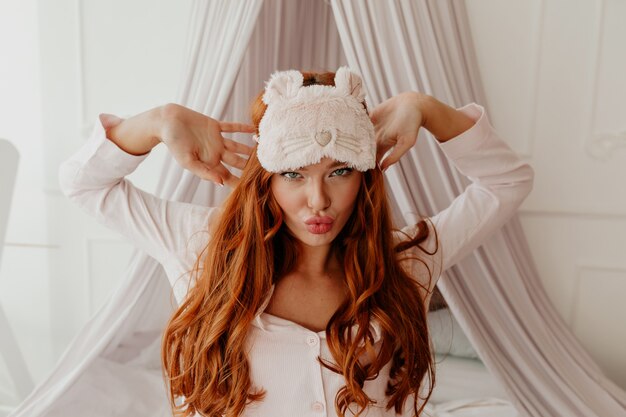 Close Up retrato de mujer divertida salido con largo cabello rojo ondulado con máscara para dormir hace muecas en la cama