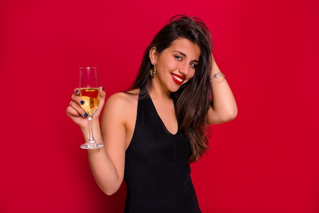 Close-up retrato de mujer feliz sonriente con cabello largo oscuro posando con una copa de champán sobre fondo rojo aislado