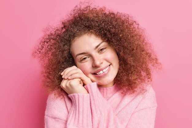 Close up retrato de mujer gentil complacida mantiene las manos cerca de la cara sonríe agradablemente inclina la cabeza mira tiernamente muestra los dientes admira algo usa poses de jersey casual en el interior contra la pared rosa