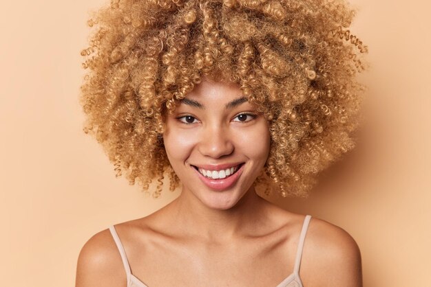 Close up retrato de mujer de pelo rizado positivo sonríe suavemente tiene piel sedosa y saludable sin maquillaje plantea hombros desnudos mira directamente a la cámara aislada sobre fondo beige. Concepto de belleza.