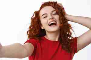 Foto gratuita close up retrato de niña de jengibre despreocupada y feliz, tocando su cabello rizado natural y riendo mientras toma selfie en la pared blanca