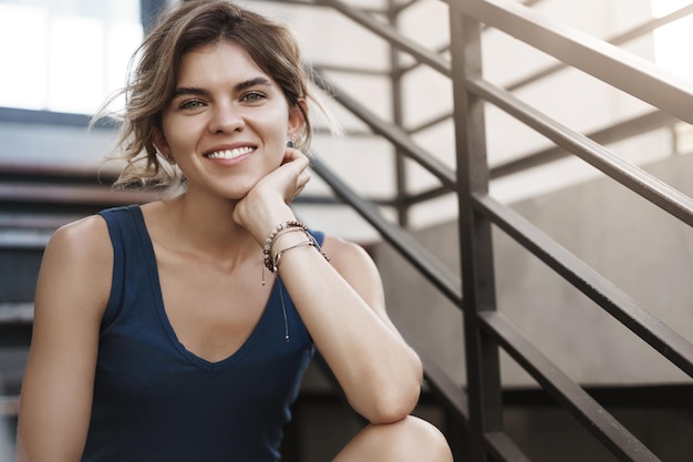 Closeup atractiva despreocupada ambiciosa joven estudiante universitaria europea escuchar buenas noticias sonriendo encantado sentado solo fuera de las escaleras relajado feliz toque mandíbula palma mirada cámara